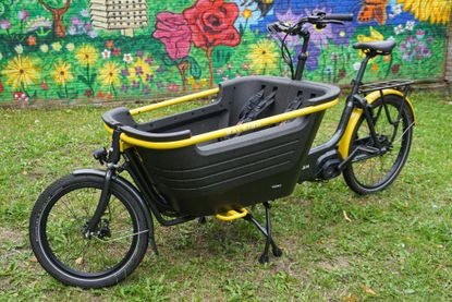 This image shows the Raleigh Stride 2 e-cargo bike on grass with a colourful wall in the background