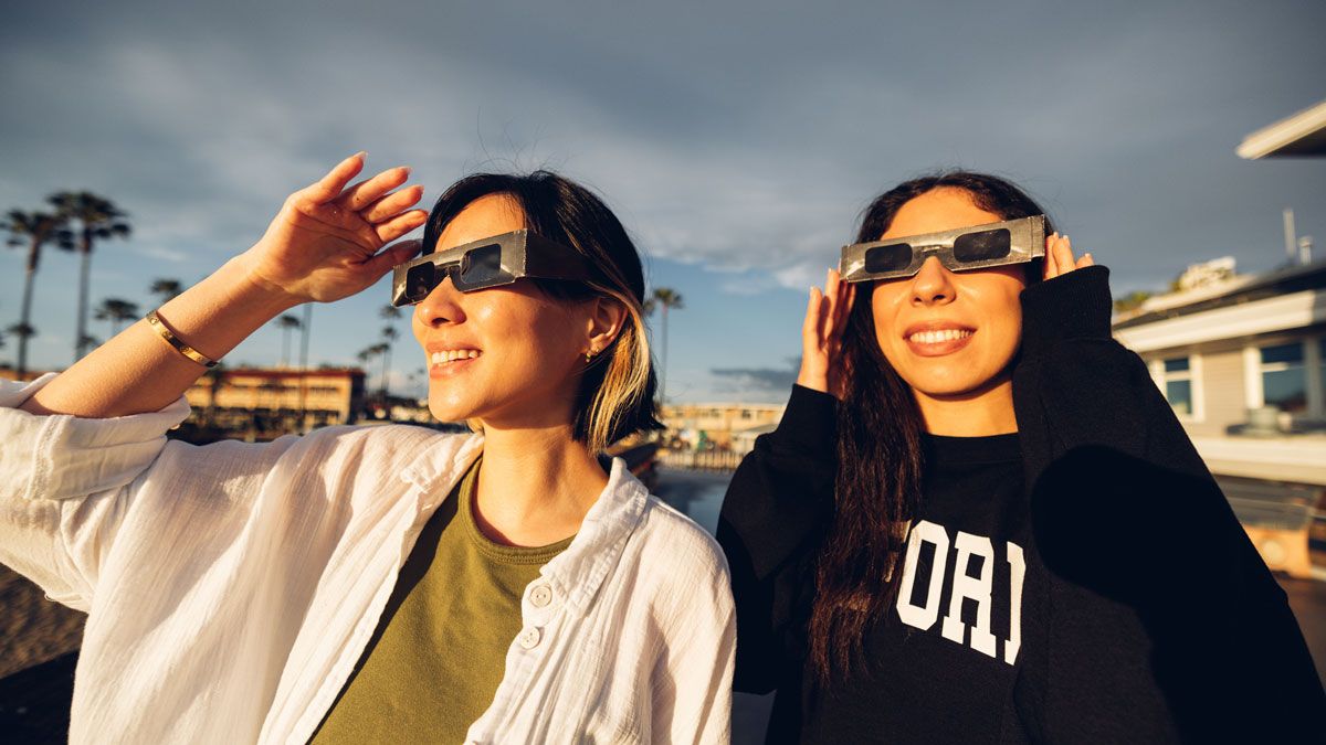 Two women wearing solar eclipse glasses while looking at the sun with palm trees in background.