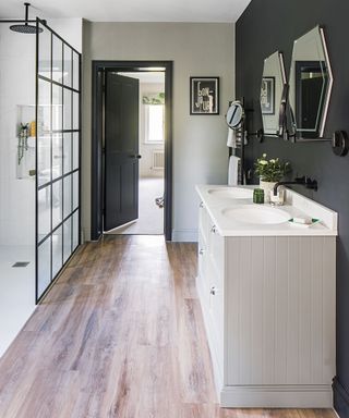 contemporary bathroom with wooden floor, black statement wall with two hexagonal mirros above a twin sink, with a large walk in shower with glass wall divider with black panels, and a black bathroo entrance door