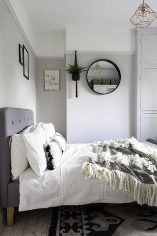 A guest bedroom with extremely light pink-purple wall colour, white bed linen, grey upholstered bed, rug and round mirror