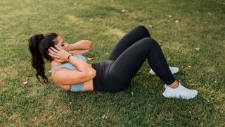 Woman performs a crunch outside on grass. lying on her back with knees bent and feet flat on the floor, and her head and shoulders raised off the floor 