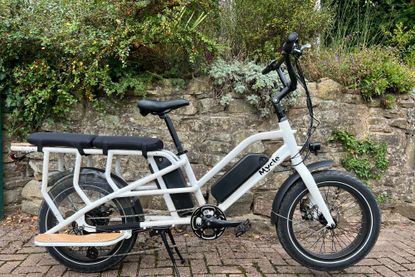 This image shows the Mycle Cargo Electric bike in full with a stone wall and greenery in the background