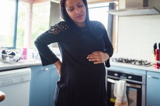 Signs of labor: A pregnant woman holds her back and belly as she braces in pain from a contraction while standing in her kitchen. 