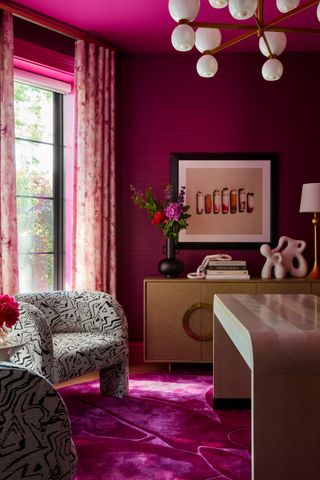 A magenta-painted, maximalist home office. There are two black and white accent chairs by the window and a magenta rug beneath them. There is a mid-century style console table with an organically shaped white vase and a similar black vase.