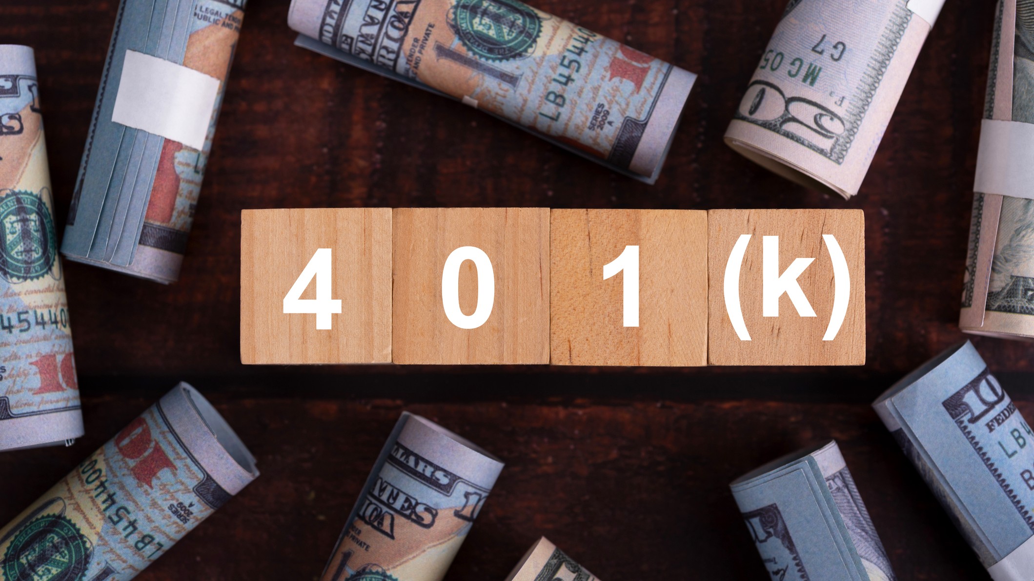 Wooden blocks spelling out "401(k)" are surrounded by rolls of $100 and $50 bills.