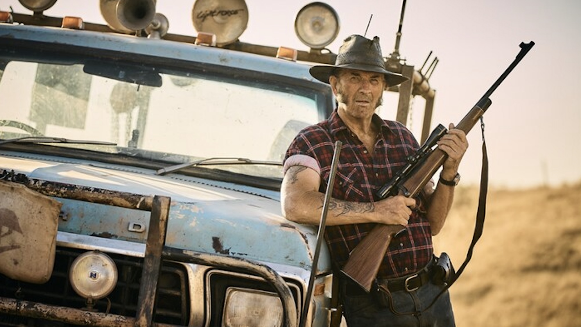 Mick Taylor leaning against truck holding a rifle in Wolf Creek Legacy