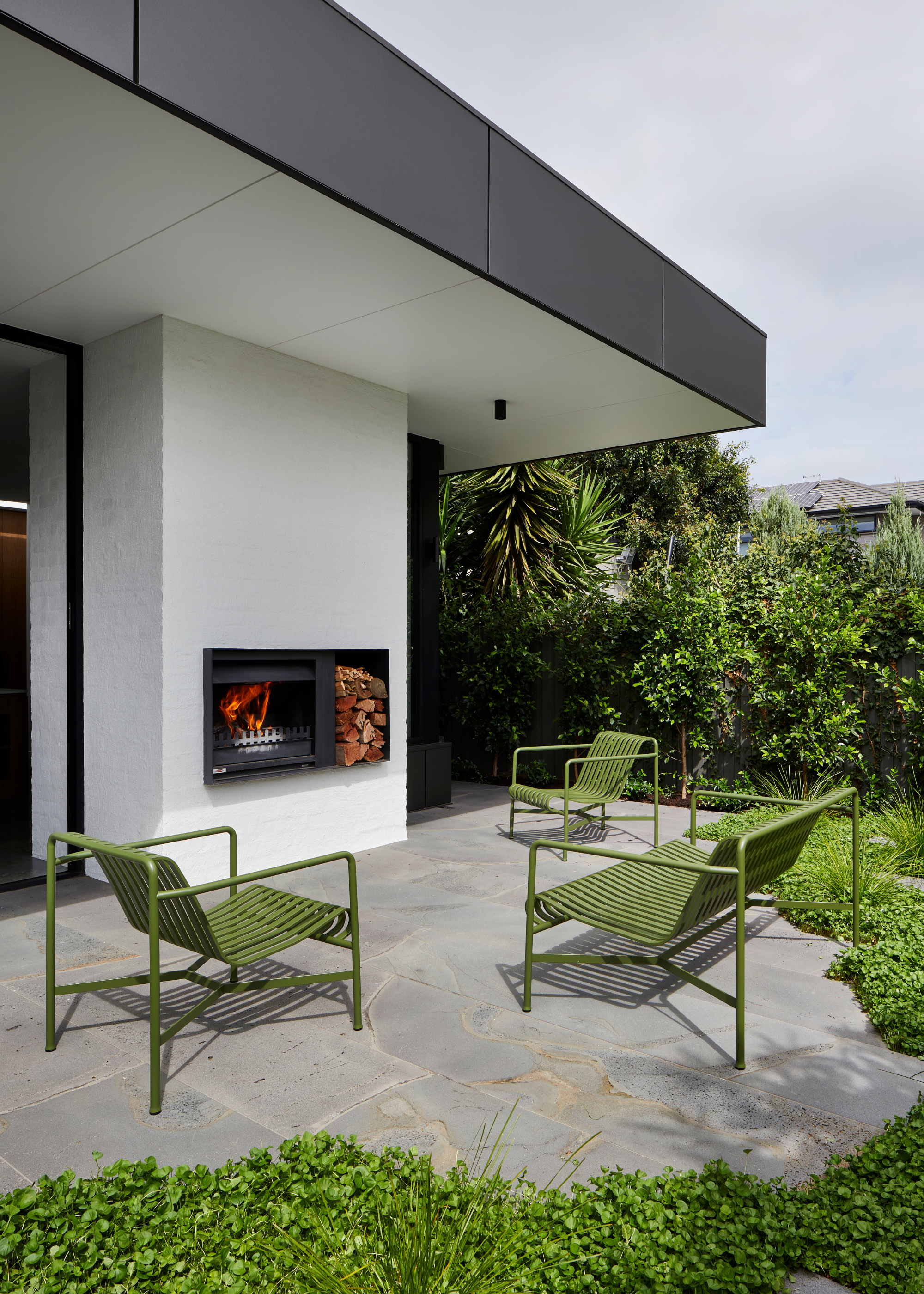 An outdoor seating space with green metal chairs neat a fireplace and lush green borders
