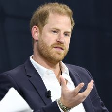 Prince Harry wears a suit with an open white shirt and gesticulates, following news he's offering continued support to a charity beloved by his late mom Princess Diana