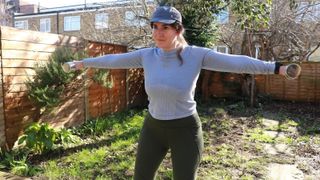 Writer Marina Leiva García works out in her garden on a sunny day. She is walking across the lawn, holding two dumbbells out at shoulder height. She wears a cap and a long-sleeved top.