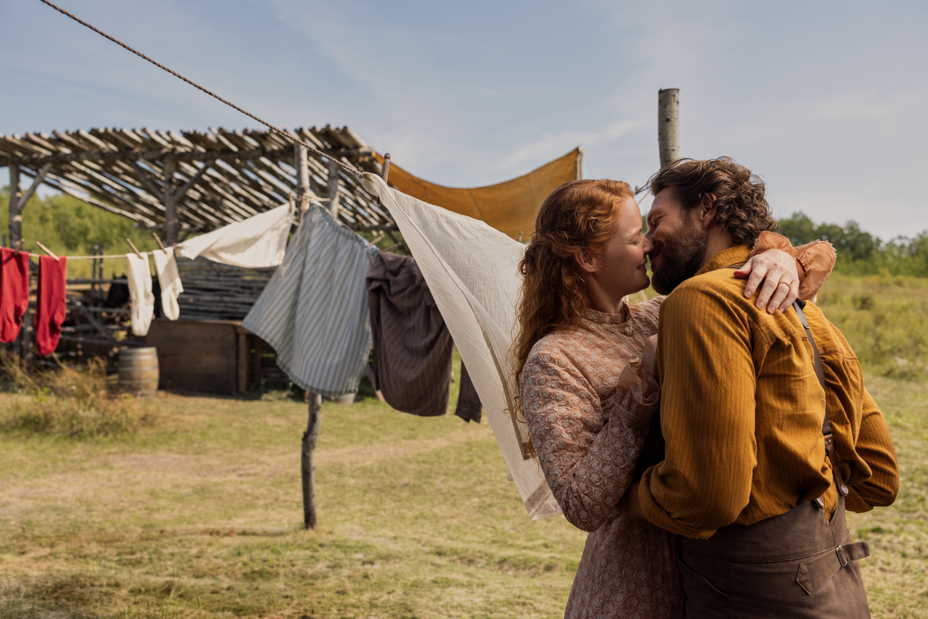 Crosby Fitzgerald as Caroline Ingalls and Luke Bracey as Charles Ingalls kiss while standing in front of a full clothesline and a wooden cabin, in Netflix's 'Little House on the Prairie.'