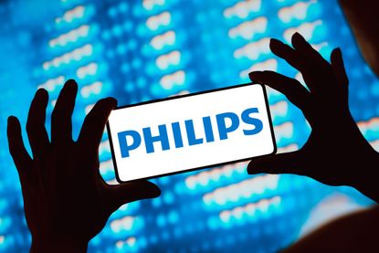 Hands hold a smartphone with the Philips logo displayed, in front of a lighted background. 