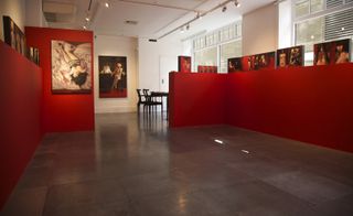 Display room, slate grey floor, white wall and ceiling with spotlights, windows letting in light right side and, red wall surround display with female erotic art along the top, black table and chairs far left, female erotic artwork on far wall