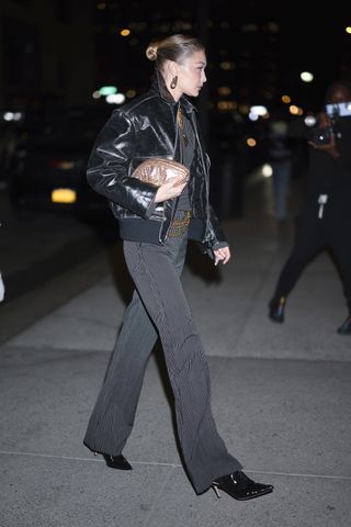 Gigi Hadid wears a leather jacket, pinstripe set, a gold clutch, black boots, and gold chain jewelry.