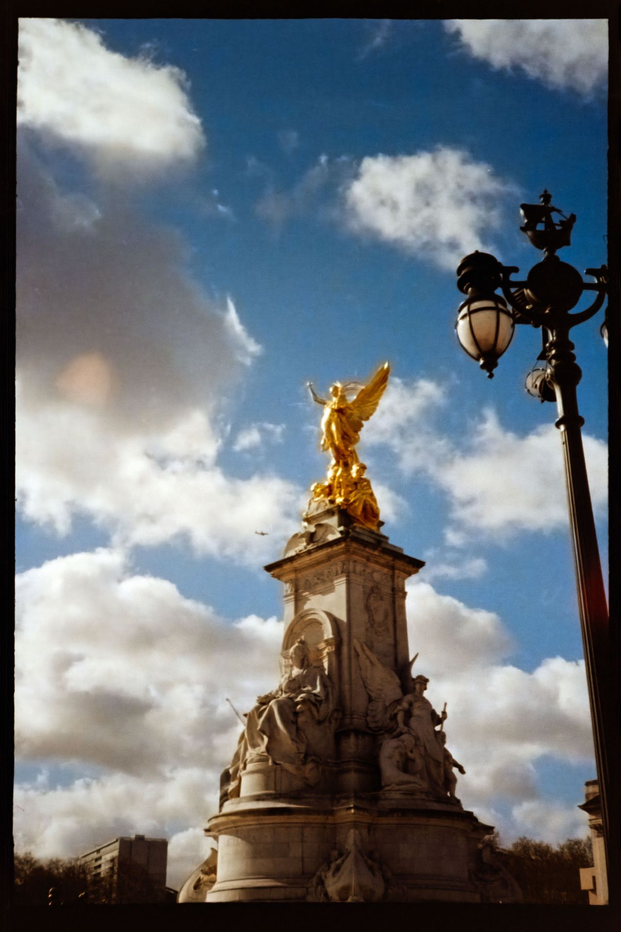 Scanned film photograph of the Victoria Memorial in London, with its golden statue framed against a blue sky and dramatic white clouds.