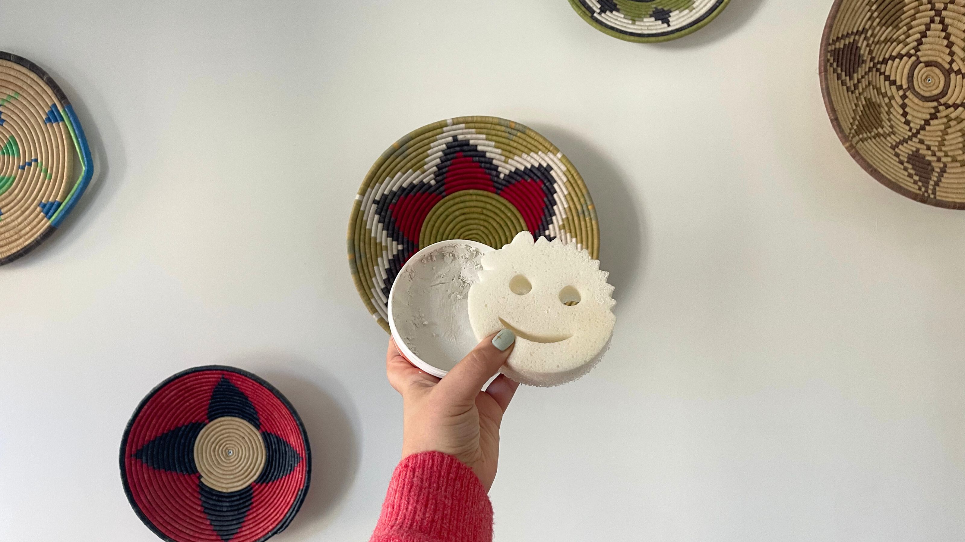 Hand holding Scrub Daddy Power Paste and sponge in front of a white wall with colourful baskets on it