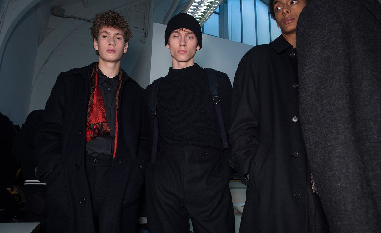 Three male models wearing looks from Margaret Howell&#039;s collection. One model is wearing a black shirt, trousers and coat with a red and black scarf. Another model is wearing a black high neck jumper, black trousers and backpack. And a third model is wearing a black, buttoned coat