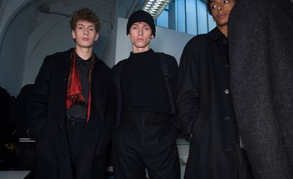 Three male models wearing looks from Margaret Howell's collection. One model is wearing a black shirt, trousers and coat with a red and black scarf. Another model is wearing a black high neck jumper, black trousers and backpack. And a third model is wearing a black, buttoned coat