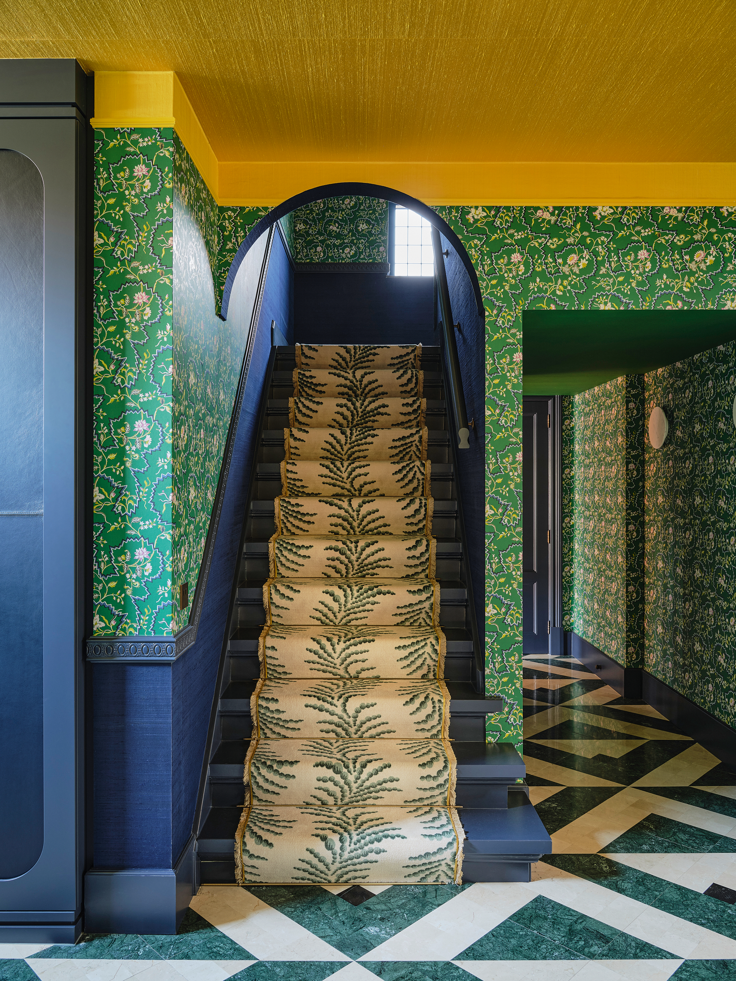 Hallway with green floral patterned wallpaper, green and white geometric marble floor, blue panelling and stairs with green and white fern runner, and yellow painted ceiling