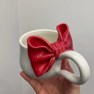 A hand holding a white coffee cup with large red bow detail above the white handle, in front of a plain white background.