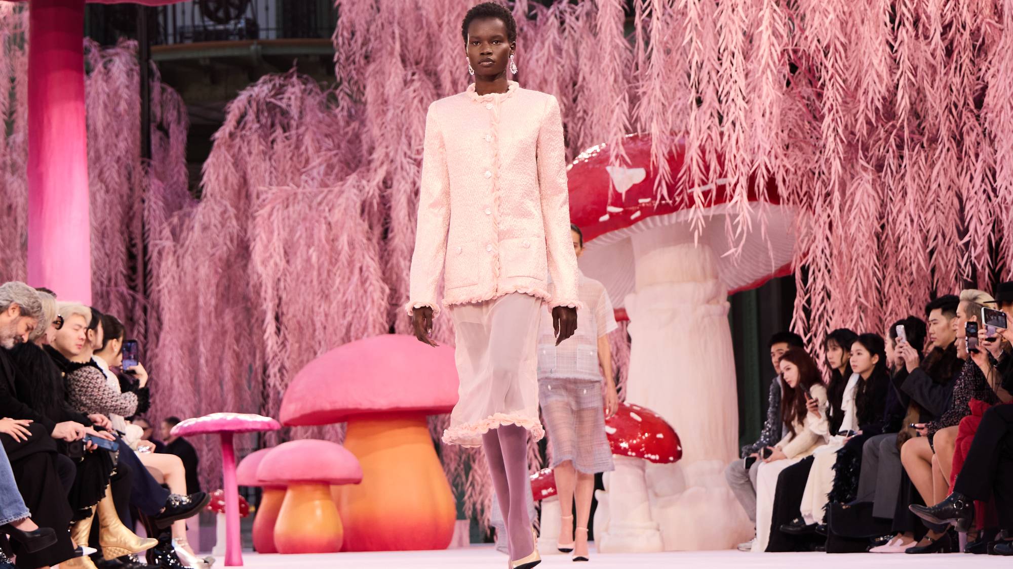 A model walks the runway surrounded by oversized mushrooms and pink leaves