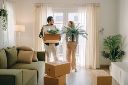 A man and a women moving into a new house, surrounded by cardboard boxes, a few plants, and a grey sofa.