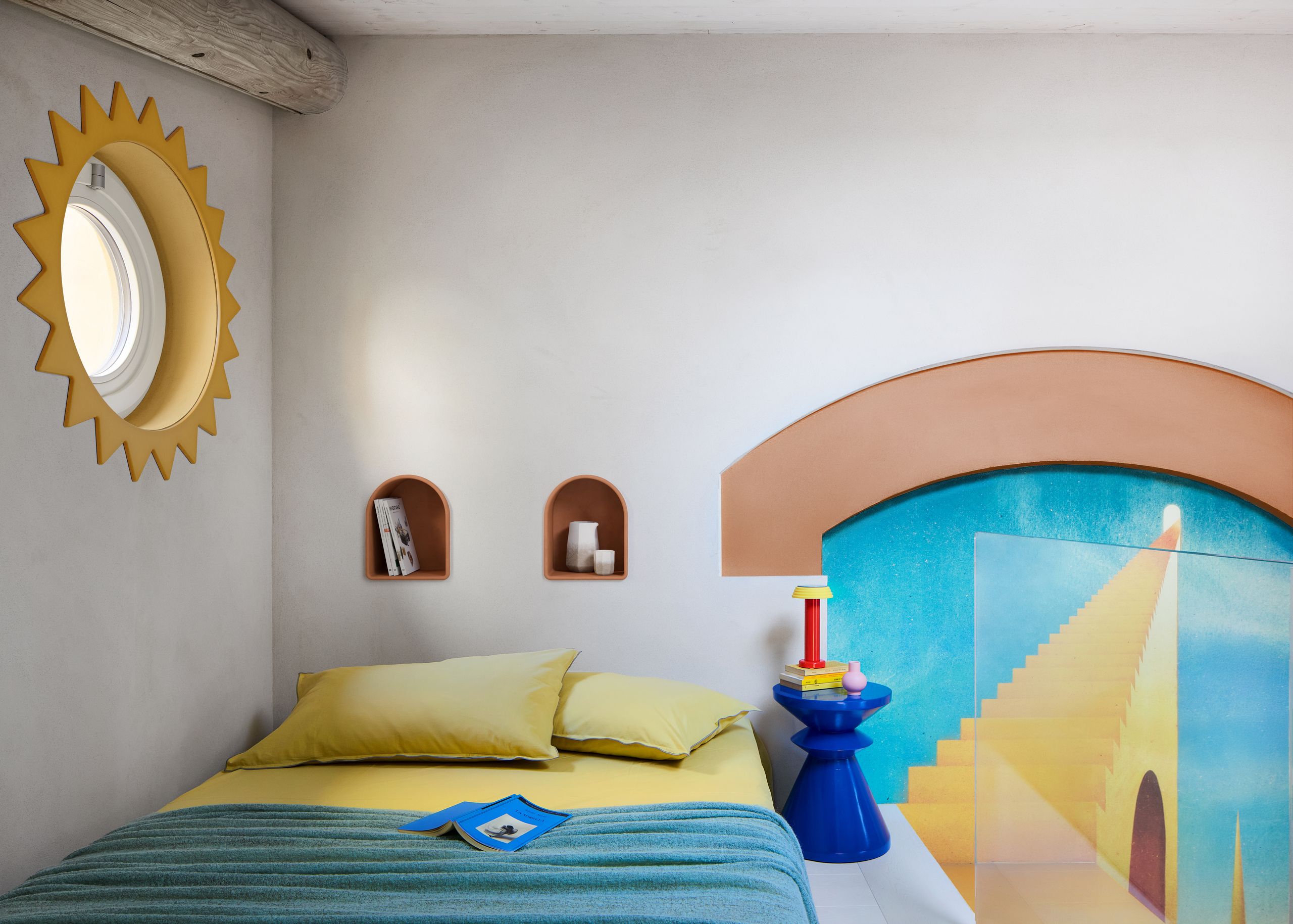 A loft bed in a white apartment with a bright mural on the wall, a blue stool, yellow bedding, a blue coverlet, arched storage nooks, and a porthole window with a sun-like border
