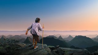 An elderly woman with a walking stick stands on a rocky ledge, gazing over majestic mountains at sunrise