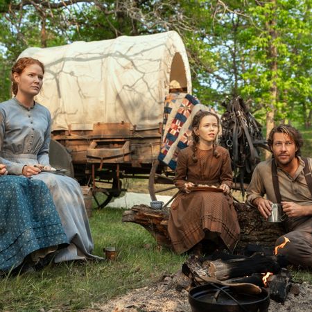 The Ingalls family (Skywalker Hughes as Mary, Crosby Fitzgerald as Caroline, Alice Halsey as Laura, Luke Bracey as Charles) sits around a woodland campsite with a covered wagon behind them, in Netflix's 'Little House on the Prairie.'