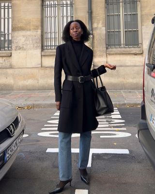 French woman poses in the street wearing a black coat, belt, straight leg jeans and kitten heels