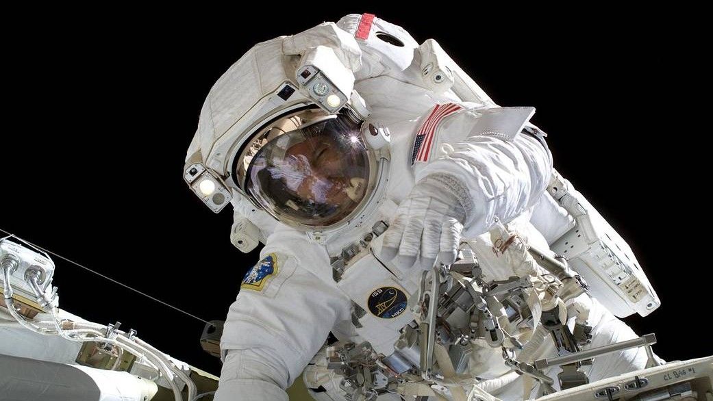 Astronaut shown doing a spacewalk outside the International Space Station. They are in the center of the image and appear to be bending towards the camera