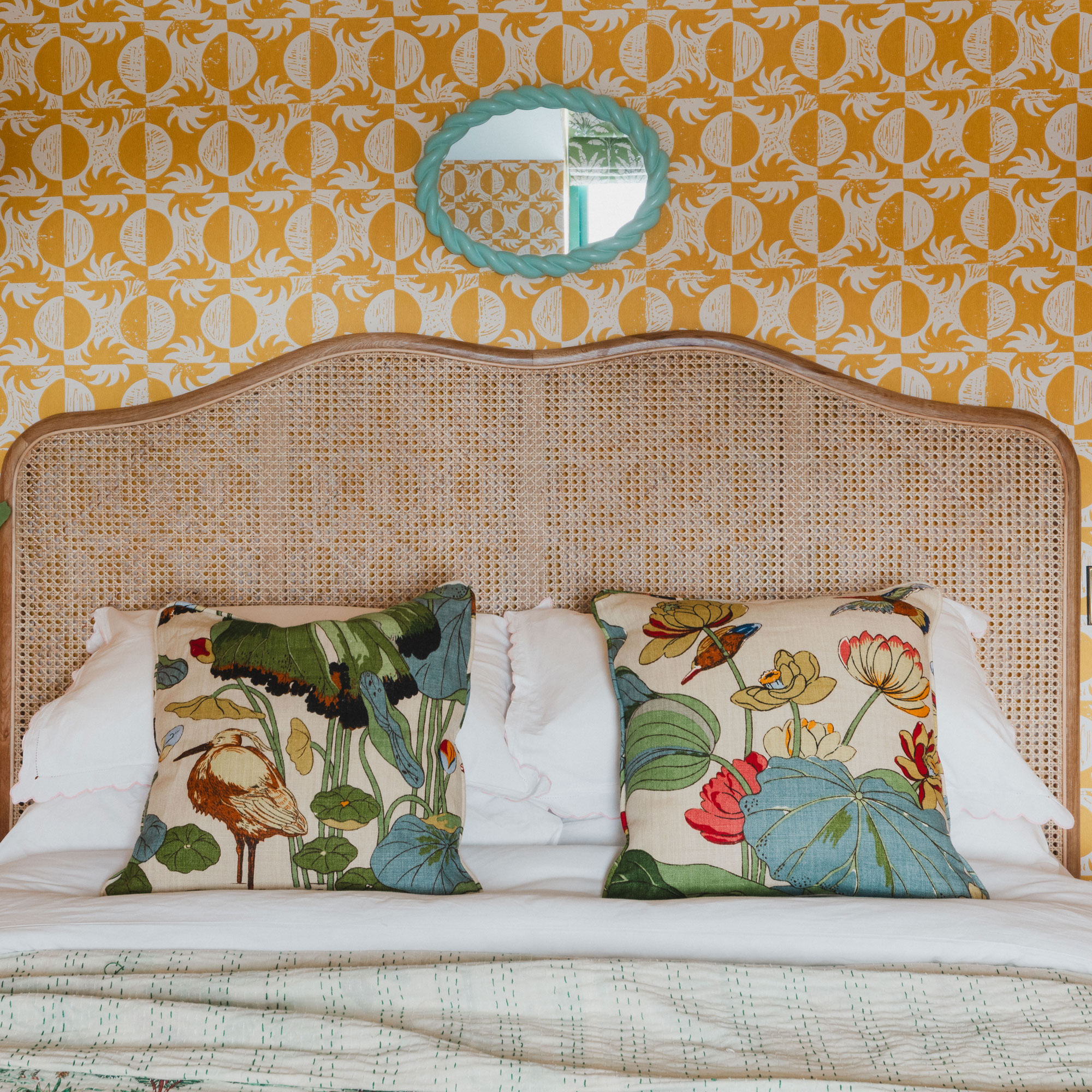 Yellow bedroom wallpaper with rattan headboard, turquoise mirror and floral throw cushions