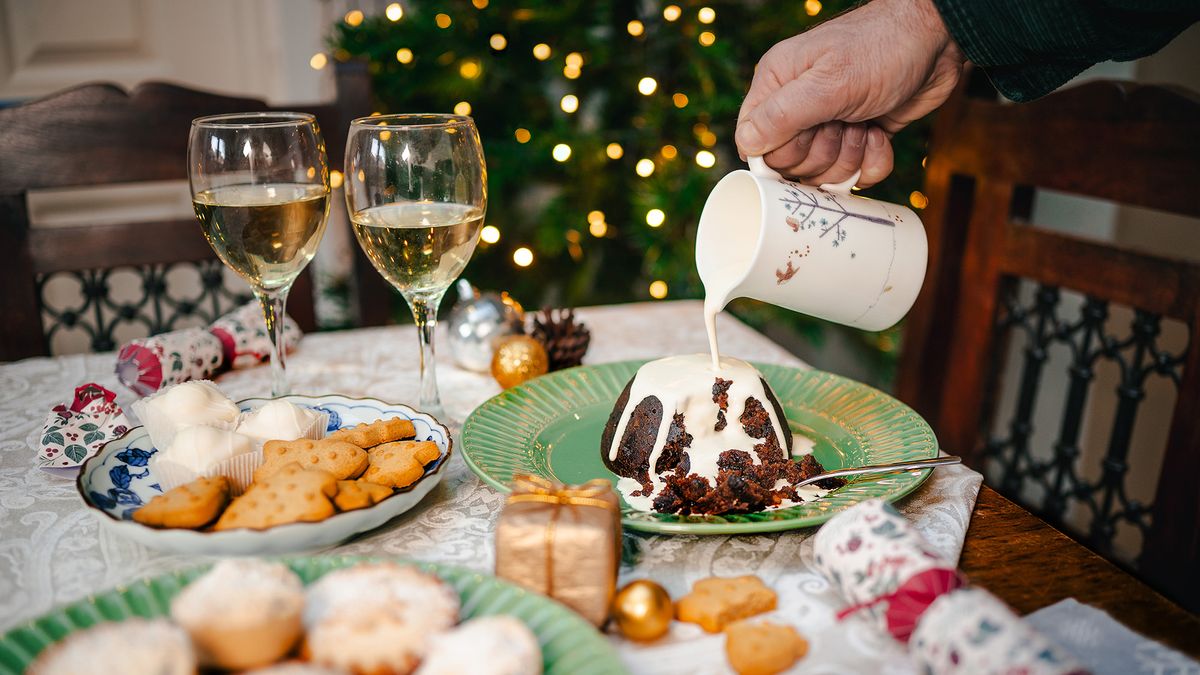 Hand holding cream and pouring it over Christmas pudding, sat on table displaying a festive spread. Bokeh fairy lights in background 