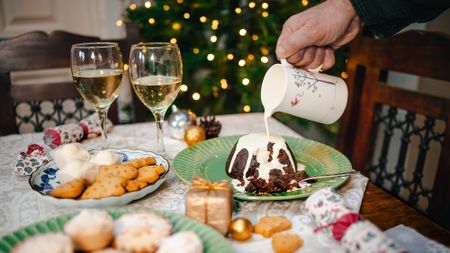 Hand holding cream and pouring it over Christmas pudding, sat on table displaying a festive spread. Bokeh fairy lights in background 