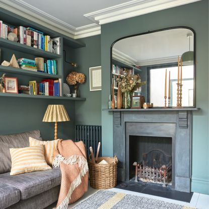 A dark green-painted living room with a black fireplace and a large mirror above and floating shelves above a grey sofa