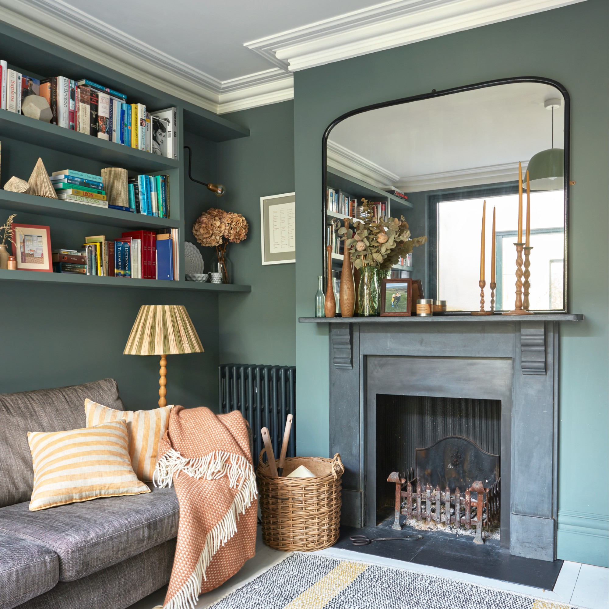 A dark green-painted living room with a black fireplace and a large mirror above and floating shelves above a grey sofa