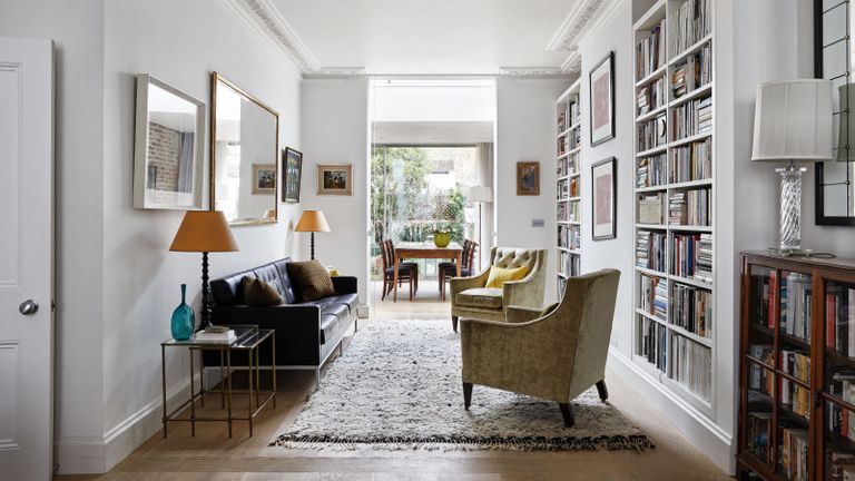 Small home library ideas. Bright open-plan living and dining space, white painted walls, floor to ceiling shelving filled with books, cozy seating area with sofa and two armchairs, soft gray textured rug underneath, lots of framed pictures and paintings on walls, reading and desk lamps beside seating areas, rectangular wall mirror above sofa