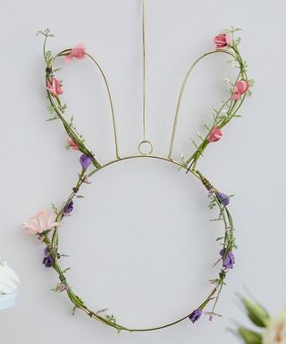 A gray wall with a bunny head shaped gold wreath hung on it with green grass and pink and purple flowers around it