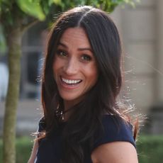 Meghan Markle has long brown hair and is smiling on the eve of her wedding while wearing a navy dress