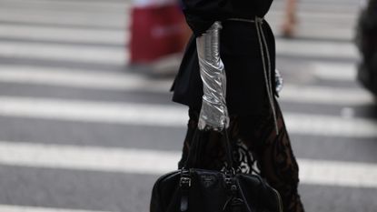 A Fashion Week guest is seen wearing big silver hoop earrings, black sunglasses from Celine, a long black blazer with a rhinestone ribbon as a belt, underneath a transparent skirt with black pattern, shiny silver gloves, a black leather bag from Prada and black leather heels from Maison Margiela outside before the Christian Siriano Show on September 08, 2023 in New York City.