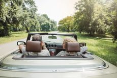A mature couple seated in a convertible car, driving down a tree-lined street.