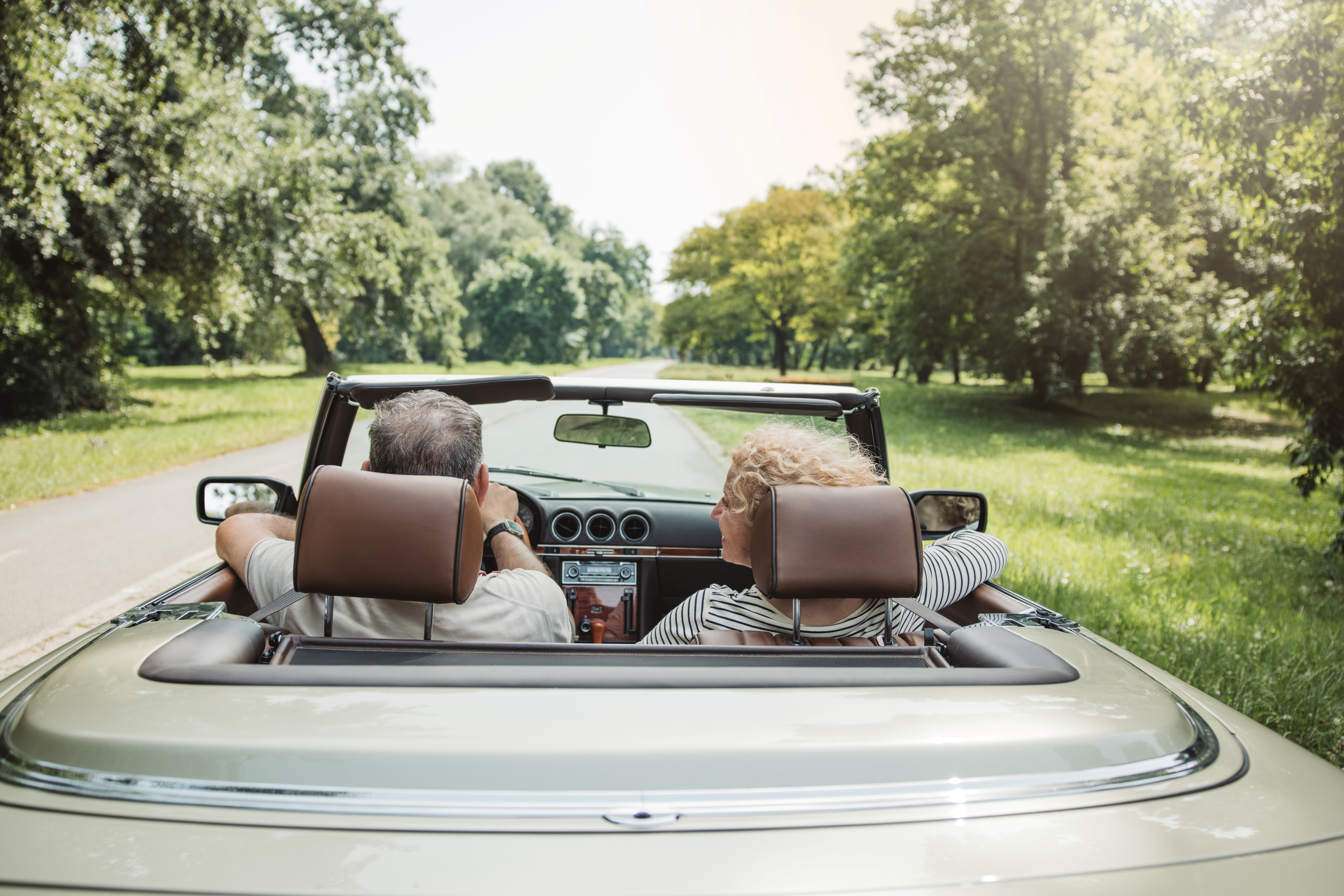 A mature couple seated in a convertible car, driving down a tree-lined street.