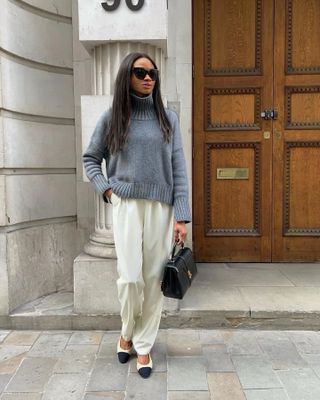 A woman wears a gray turtleneck sweater, silk pants, ballet flats, and a black bag.