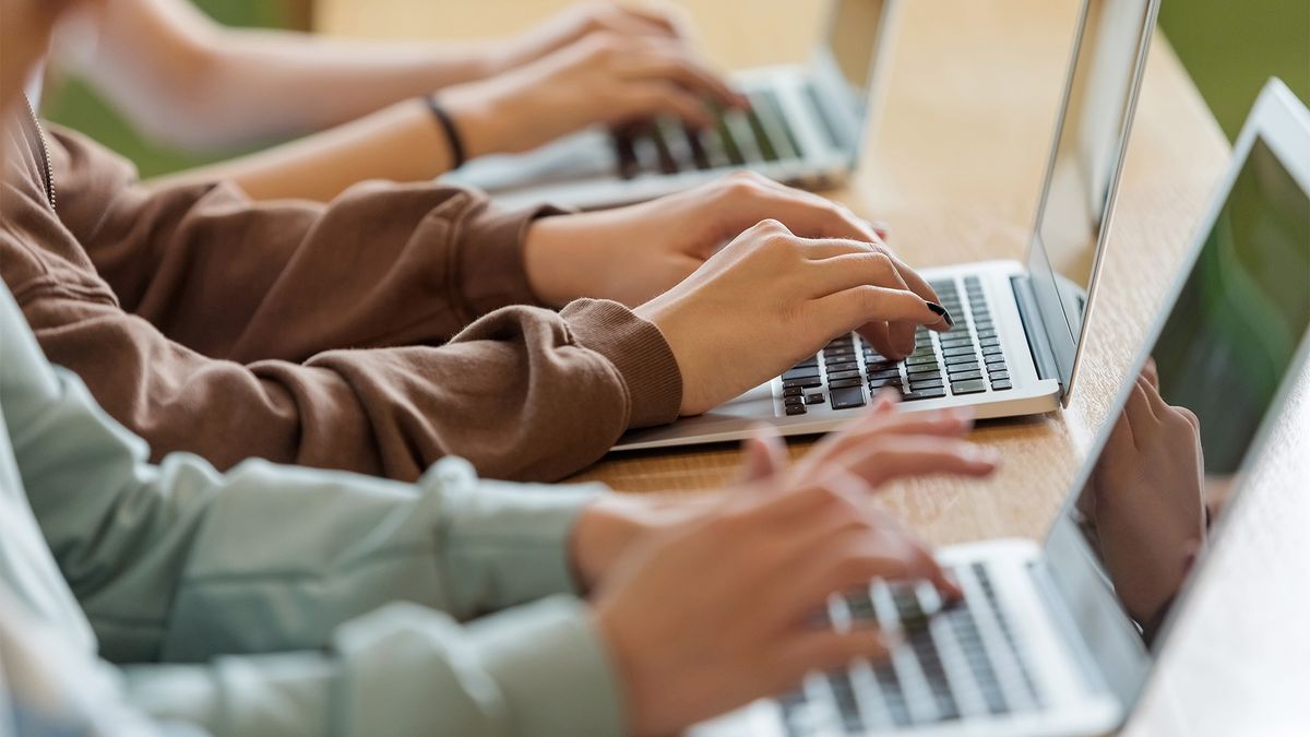 Three teenage schoolgirls typing on laptop keyboards while sitting in classroom.