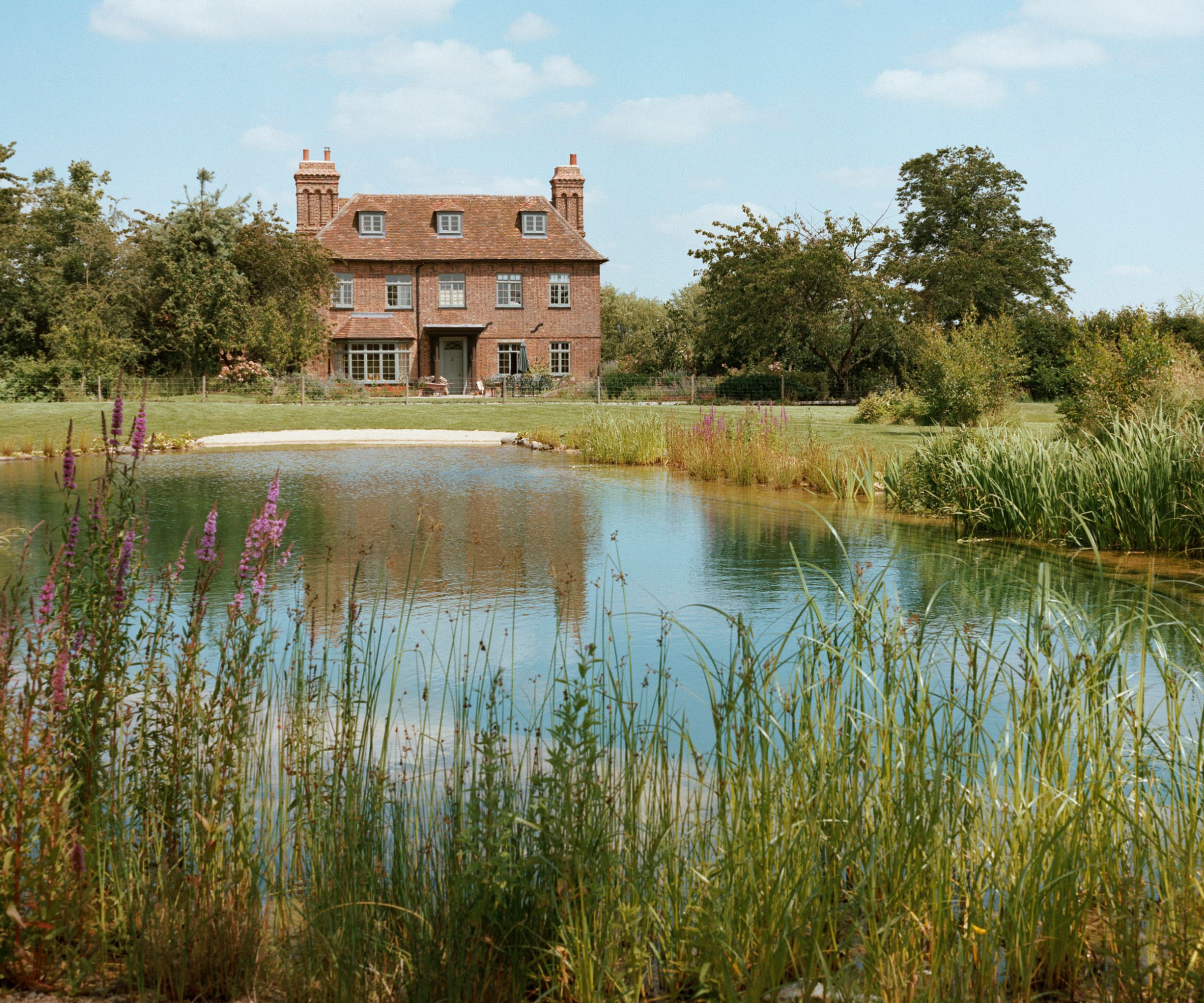 Natural swimming pool in front of a traditional red-brick house, with meadow-style planting and long grass softening the edges of the water.