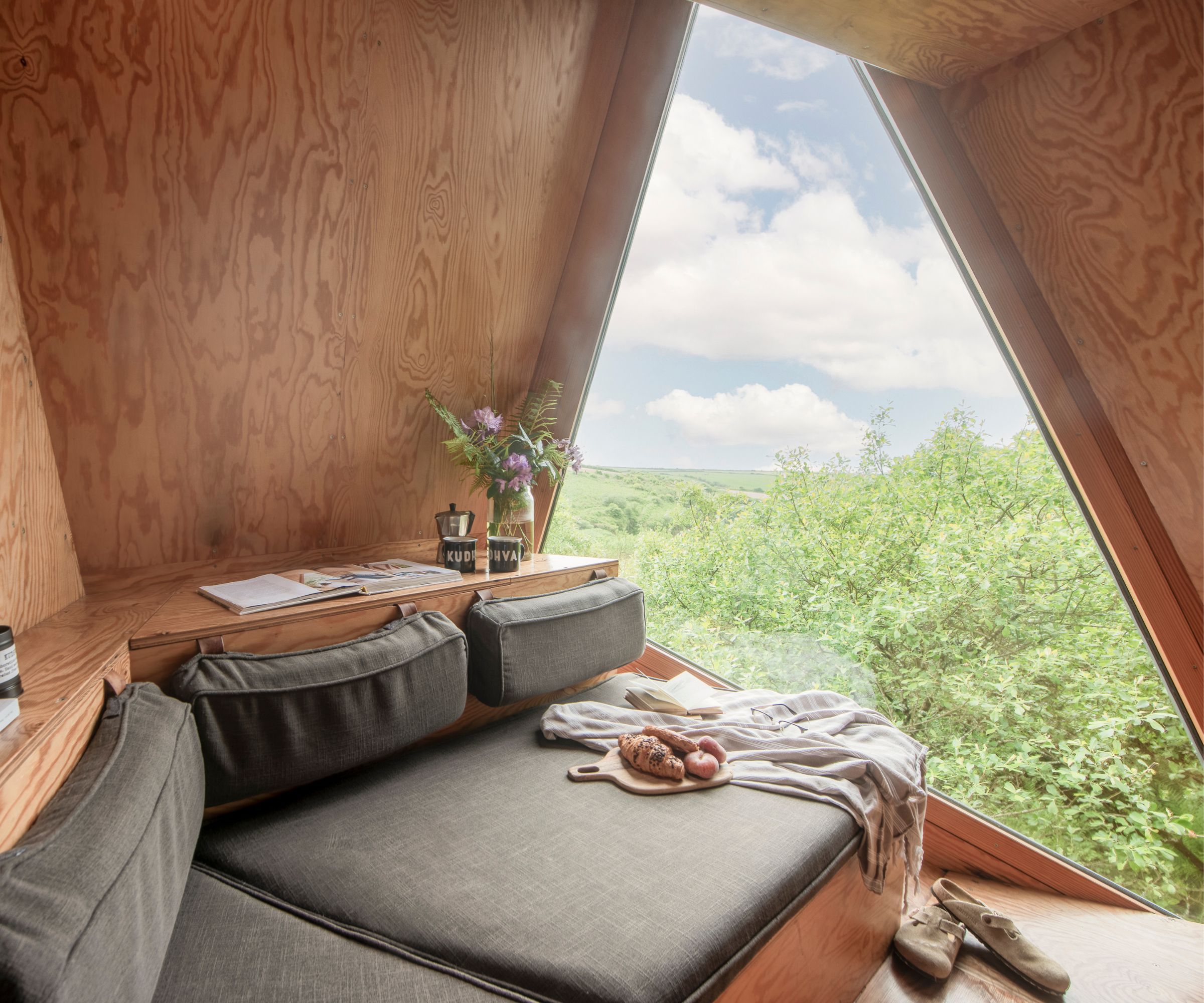 The interior of a modern wooden treehouse with a triangular window offering scenic countryside views, a built-in cushioned seating area, and a small wooden desk.