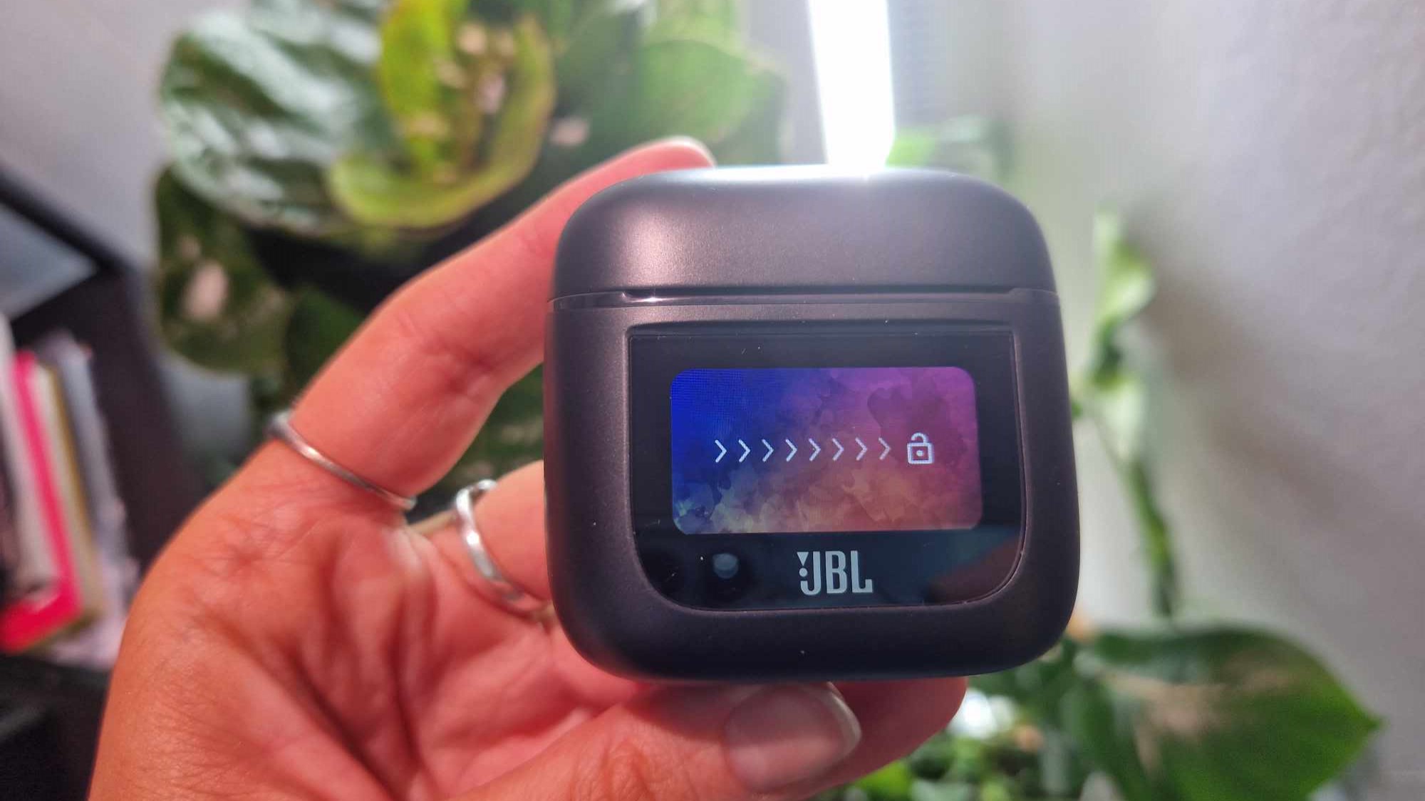 En hand håller upp fodralet till JBL Tour Pro 2, som visar dess låsskärm.