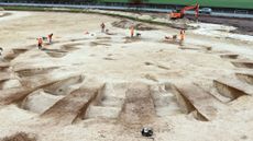 We see a dirt field with a flattened barrow (mound) that has cut marks in it like rays from a sun. There are workers and an excavator.