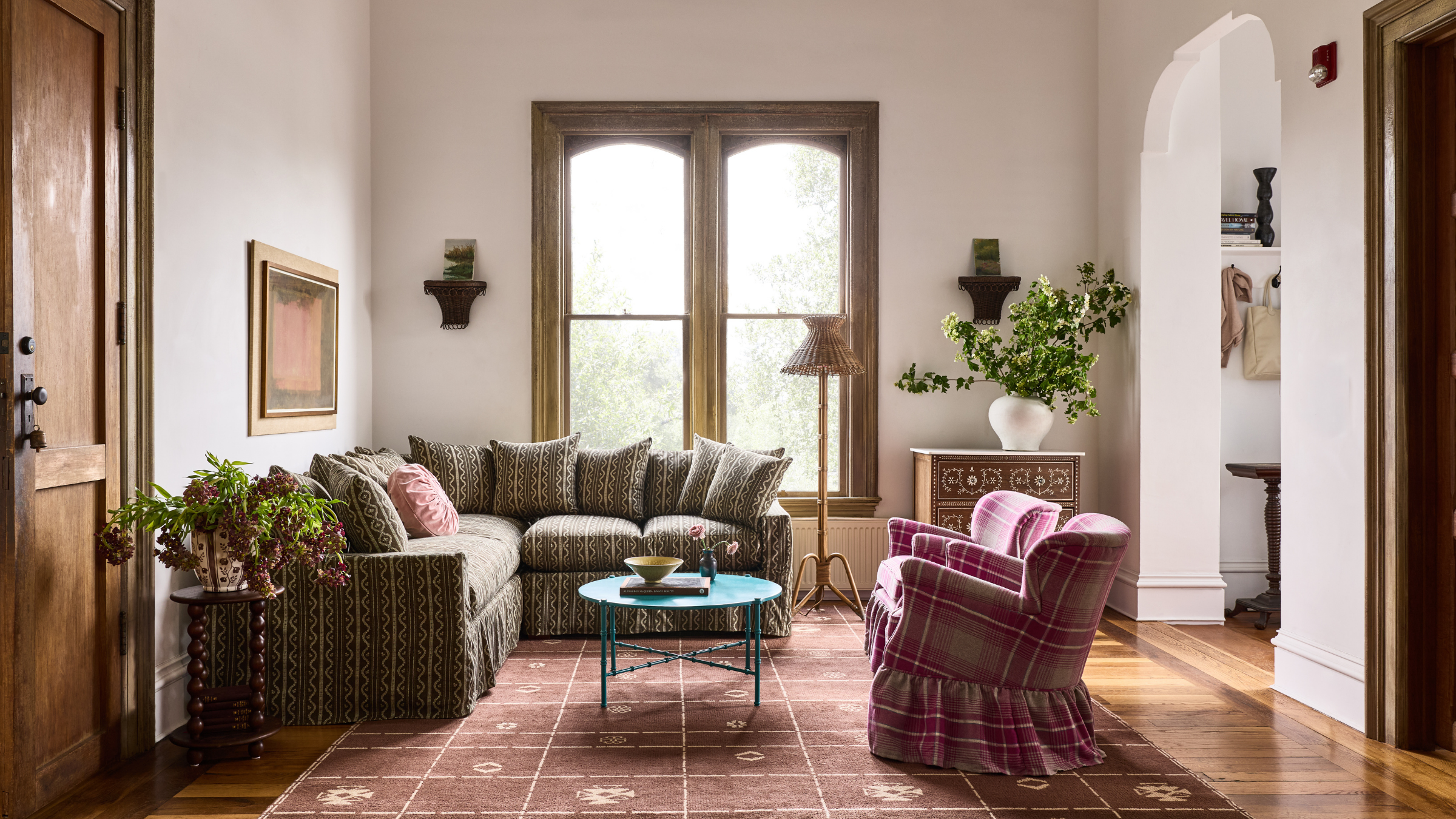 Botanically patterned sectional sofa pictured in a characterful living room with a geometric pattern rug, gingham skirted accent chair, and large, wood-framed windows