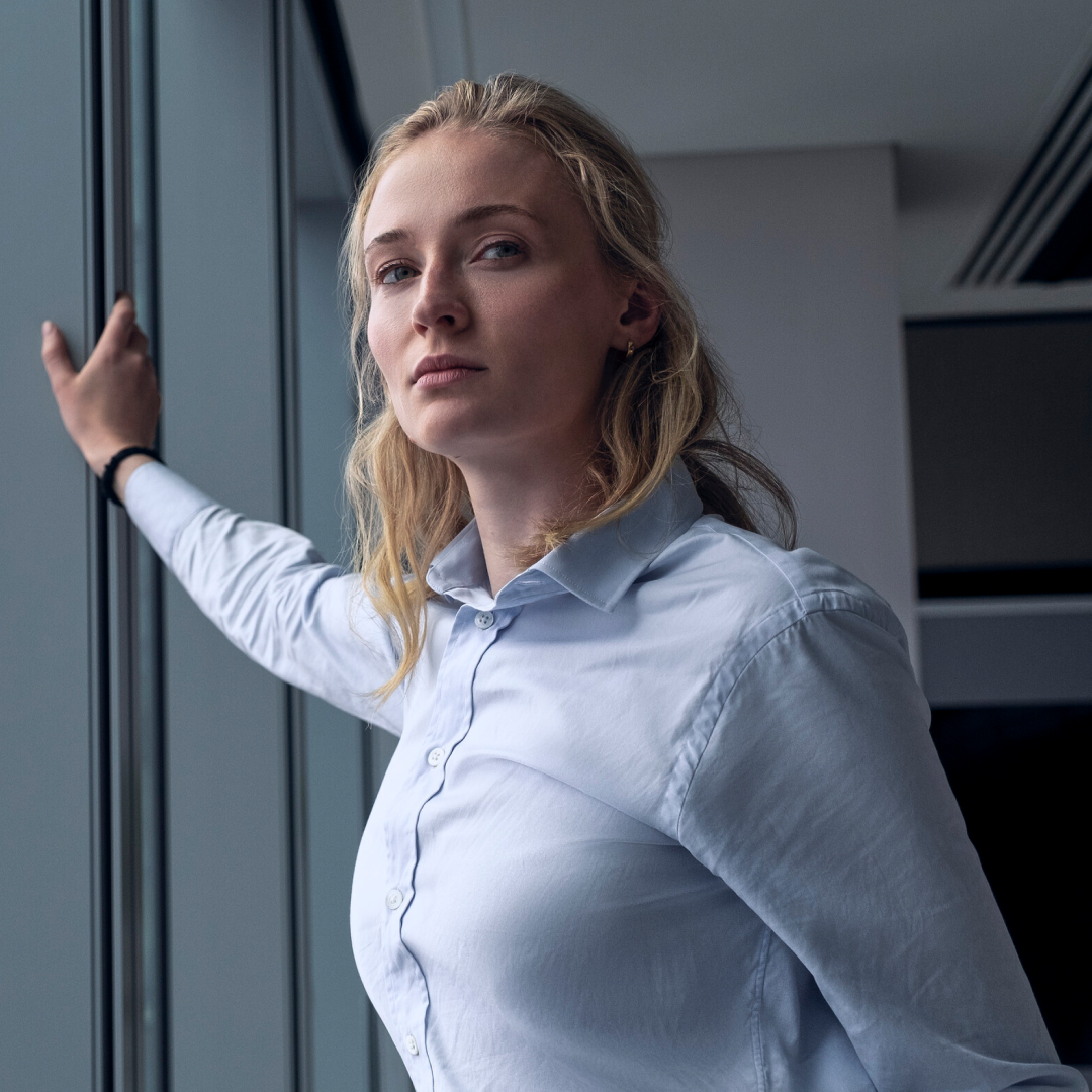 sophie turner as zara wearing a light blue button down leaning against a window in an office building in a still from steal