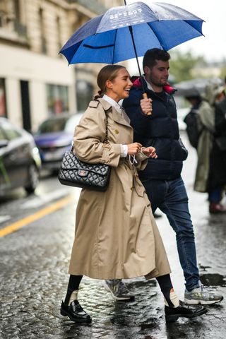 A woman wearing a tan trench coat and smiling while a man holds her umbrealla.